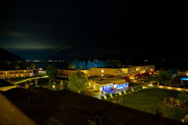 Moraitika, Yunanistan - 09: 25 2022: Yüzme havuzlu otelin avlusunun aydınlatılmasıyla akşam manzarası. Gökyüzünde, uçak ışıklarından oluşan bir iz. Uzun pozlama fotoğrafı. Yüksek kalite fotoğraf