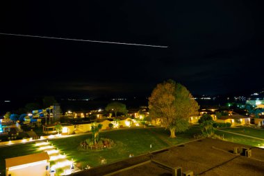 Moraitika, Yunanistan - 09: 25 2022: Yüzme havuzlu otelin avlusunun aydınlatılmasıyla akşam manzarası. Gökyüzünde, uçak ışıklarından oluşan bir iz. Uzun pozlama fotoğrafı. Yüksek kalite fotoğraf