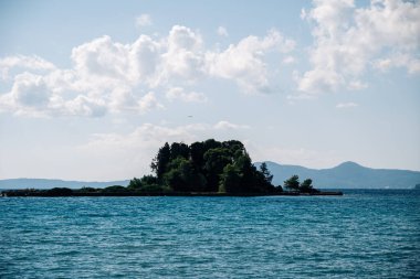 Küçük bir ada, kümülüs bulutları ve ufukta dağlarla ve kıyı arazileriyle çevrili engin bir su kütlesinin ortasında huzur içinde oturmaktadır.