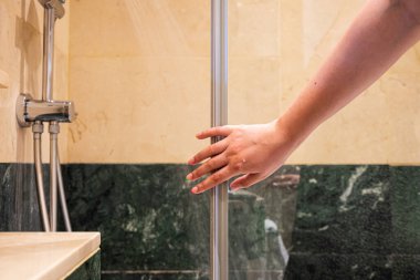 Parmakları açık bir insan eli cam duş kapısına doğru hareket eder ve açmaya hazırdır. Bilek ve başparmak ahşap çerçeveye yerleştirilmiş, arka planda bel var.