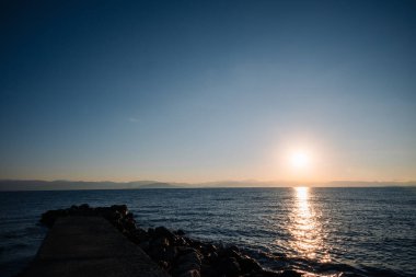 Okyanus sakin ve gökyüzü mavi. Güneş batıyor ve su ışığı yansıtıyor.