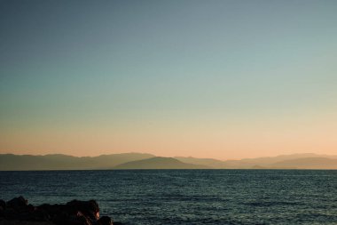Gökyüzü mavi ve okyanus sakin. Ufuk görünür, dağlar uzak.