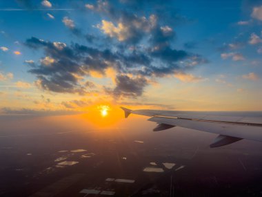 The sun sets dramatically on the horizon as seen from an airplane wing, casting warm colors over the vast fields and water bodies below while clouds scatter the sky. clipart