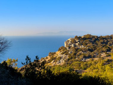 Berrak sular ufka kadar uzanıyor. Kayalık kayalıklar ve yemyeşil alanlar ılık güneşin altında sakin bir kıyı manzarası oluşturuyor..