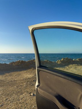 Açık araba kapısı gün batımında okyanus ve gökyüzünün çarpıcı bir görüntüsünü ortaya çıkarıyor. Sıcak ışık huzurlu bir ortam yaratırken dalgalar yavaşça kayalara çarpıyor..