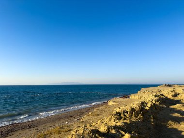 Huzurlu bir sahil manzarası, ufukta batan güneşin yumuşak ışığını yansıtan açık mavi gökyüzü ile nazik dalgaların üzerinde yükselen altın uçurumları sergiliyor..