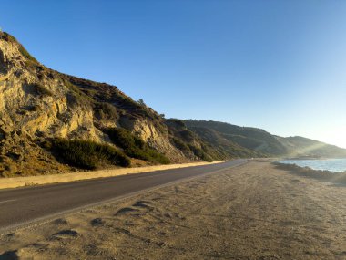 Huzurlu bir sahil yolu, öğleden sonra güneş batmaya başladığında tepelerle çevrili su boyunca uzanır ve sakin ve sakin bir atmosfer yaratır..