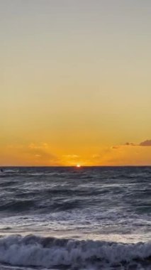 Güneş ufukta batarken turuncu ve sarı muhteşem renkler gökyüzünü dolduruyor. Hafif okyanus dalgaları kıyıya vurur ve alacakaranlıkta huzurlu bir atmosfer yaratır..