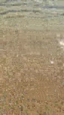 Yumuşak dalgalar altın kumda dingin desenler oluşturur. Güneş ışığı berrak sulardan yansıyarak güneşli bir yaz günü boyunca sakin bir sahil ortamını ortaya çıkarır..