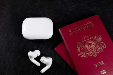 Jurmala, Letonya - 01.05.2025: Apple Kulaklıklar - Airpod Pro 2. Kablosuz kulaklıklar, karanlık bir yüzeyde açık bir pasaportun yanında oturur ve gerekli seyahat ekipmanlarını ön plana çıkarır.. 