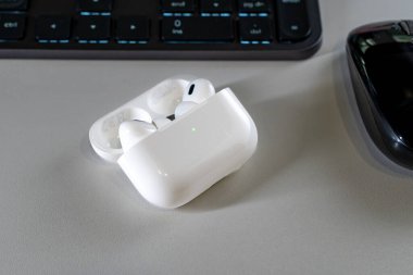 Jurmala, Letonya - 01.05.2025: Apple Kulaklıklar - Airpod Pro 2. Kablosuz kulaklıklar şarj masasının üzerinde duruyor. Modern bir ofis ortamında siyah bilgisayar faresi ve klavyesi yakınlarda görünür..