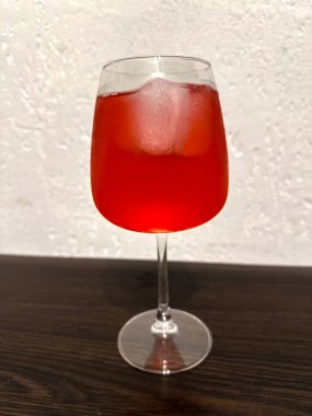 Canlı kırmızı bir kokteyl zarif bir şekilde buzla dolu berrak bir bardakta, zengin rengini ve davetkar görünümünü sergiliyor. İçki koyu, ahşap bir yüzeye konur. Sıcak bir ortam yaratır..