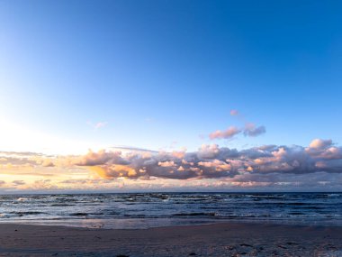 Canlı bir günbatımı okyanus dalgaları üzerinde ılık renkler oluştururken yumuşak bulutlar akşam gökyüzünde sürükleniyor. Huzurlu sahil manzarası rahatlamaya ve yansımaya davetiye çıkarıyor..