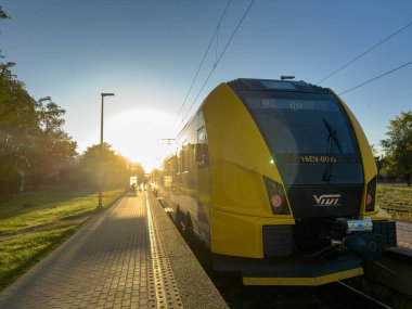 Jurmala, Letonya 14.08.2024: VİVI treni sabah Sloka tren istasyonunda. Sarı bir tren sabahın erken saatlerinde bir istasyonda duruyor. Güneş arka plandan yükseliyor, platforma ve ağaçların çevresine sıcak bir ışık saçıyor..