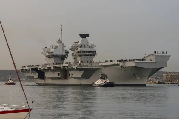 stock image Portsmouth, England, UK. 8 September 2023.  HMS Queen Elizabeth underway leaving Portsmouth dockyard at dusk with an escort of tugs and security vessels. Naval personnel line the decks.