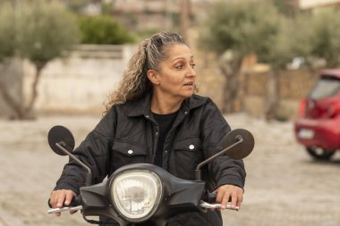 Malia, Girit, Yunanistan. 03.10.2023. Kıvırcık saçlı, parlak ceketli, motosikletli Girit bir kadın..