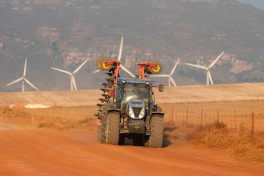 Gouda, Western Cape, South Africa. 24 April 2024.  Day end and a farm tractor makes it way on a red dirt road with a background of a Gouda wind farm and mountains. clipart