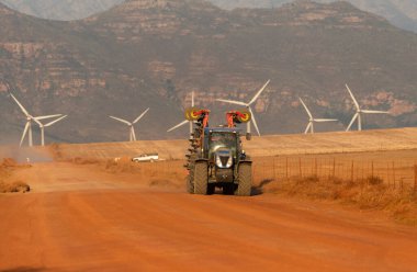 Gouda, Western Cape, South Africa. 24 April 2024.  Day end and a farm tractor makes it way on a red dirt road with a background of a Gouda wind farm and mountains. clipart