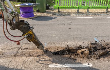 Hampshire İngiltere. 07.08.2025. Hidrolik kırıcı eklentisi, tam fiber ağ kablo kurulumu için siper kazma operasyonundan önce yol yüzeyini kırıyor. İngiltere