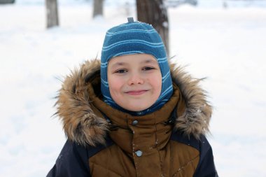 Kışın açık havada gezen çekici bir çocuğun portresi. O kalın bir şapka ve ceket giyiyor, kameraya bakıyor ve gülümsüyor. Kardaki ağaçların arka planında.