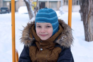 Kışın açık havada gezen çekici bir çocuğun portresi. Sıcak bir şapka ve ceket giymiş, salıncakta oturuyor, kameraya bakıyor ve gülümsüyor. Arkaplanda kar yığınları ve ağaçlar.