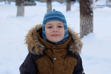 Kışın açık havada gezen çekici bir çocuğun portresi. O kalın bir şapka ve ceket giyiyor, kameraya bakıyor ve gülümsüyor. Kar yağışı ve ağaçların arka planında.