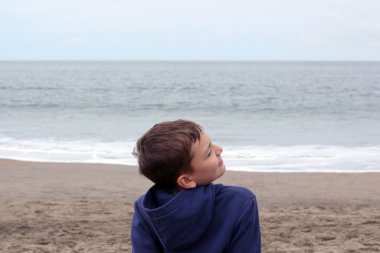 Bulutlu bir günde kumlu sahilde 10 yaşında yakışıklı sarışın bir çocuğun portresi. Oğlan sıcak bir kazak giydi. Sırtı bize dönük oturuyor..