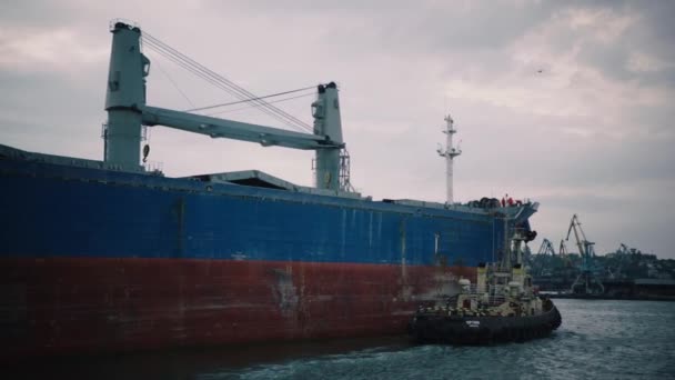 停泊拖轮的大型货船停泊在一个商业港口卸货 一条船把船向侧面推 帮助船转向 — 图库视频影像