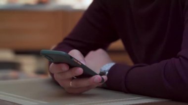 Masada akıllı telefonu tutan bir adam. Teknoloji kullanımını içeren modern, günlük bir durum. Bulanık arkaplan rahat bir kafe ya da çalışma alanını işaret ediyor.