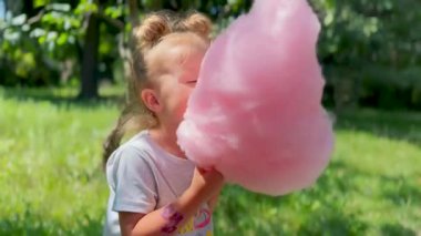 Parkta elinde büyük pembe pamuk şeker tutan ve neşeyle yemek yiyen bir kız. Çocuklar için eğlence, tatil ve yaz tatili kavramı