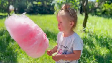 Küçük kız dışarıda pembe pamuk şeker tutuyor ve yiyor. Neşenin sembolü, çocukluk mutluluğu ve şenlik havası.