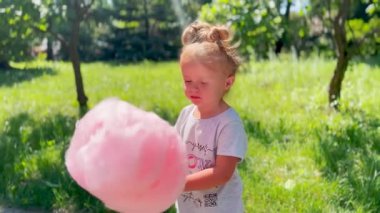 Küçük kız dışarıda pembe pamuk şeker tutuyor ve yiyor. Neşenin sembolü, çocukluk mutluluğu ve şenlik havası.