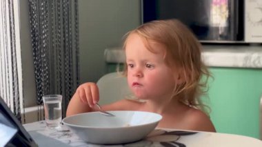 Küçük kız tablette çizgi film izlerken masada oturup lapa yiyor. Aletlerle modern çocukluk