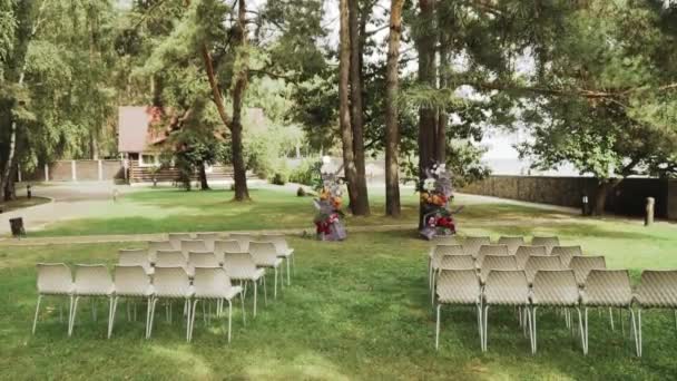 松の木の下で結婚式の花の装飾されたアーチと白い椅子のフロントビュー 遅い動き 晴れた日に公園での結婚式の外 フルHd映像 — ストック動画