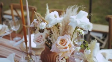 Güzel bir buket gül ve kurutulmuş çiçekleri masanın üzerine koyun ve mumlarla süsleyin. Boho tarzı düğün yemeği, tabaklar ve şarap kadehleri, vurulan kimse yok, ağır çekim. Yüksek kalite 4k