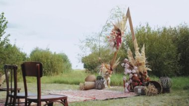 Açık hava düğün töreninde çok güzel bir bohem kemeri süsü. Pastel renklerden, kurumuş çiçeklerden, tahta sandalyelerden ve eski moda halılardan oluşan çiçek kompozisyonları. Yaz düğünü. Yüksek kalite 4k