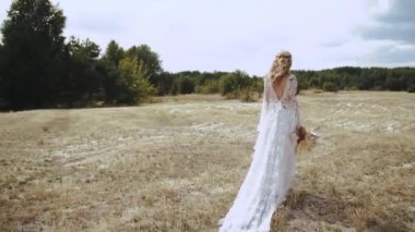 Bride in wedding boho dress walks through the field with a bouquet slow motion shot. The bride walks forward across the field and turns to the camera. High quality 4k footage