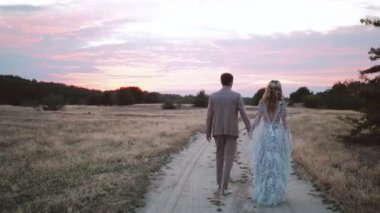 Back view of bride and groom walk through field at sunset, pink colored couds in the sky. Bride in boho style dress. Loving couple on sunset. High quality 4k footage