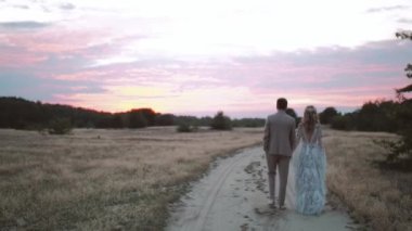 Back view of bride and groom walk through field of dried grass at sunset, pink colored clouds in the sky. Bride in boho style dress. Loving couple on sunset. High quality 4k footage