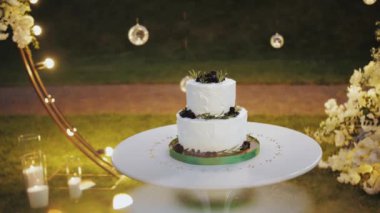 Beautiful wedding cake decorated with blueberrys in the evening near the arch, bulb lamps on the background, slow motion. High quality 4k footage