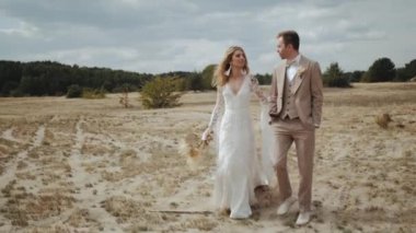 Newlyweds walk through the countryside, Bride in boho dress and groom in suit goes across the field, slow motion. High quality 4k footage
