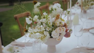 Yemek masasında güzel bir buket, yakın plan pastel çiçekler, el yapımı yeşillikler, şenlikli tabaklar ve şarap bardakları. Çiçek kompozisyonu masada, ağır çekimde. 4k görüntü