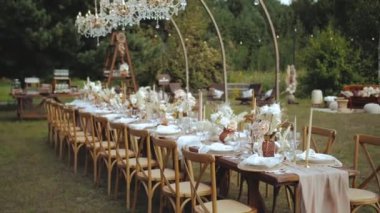 Dışarıda mumlarla süslenmiş düğün masası ve boho tarzı düğün yemeği için kurutulmuş pastel çiçekler, tabaklar ve şarap bardakları, rüzgarlar masa örtüsü, peçeteler ve çiçeklerle süslenmiş. 4k