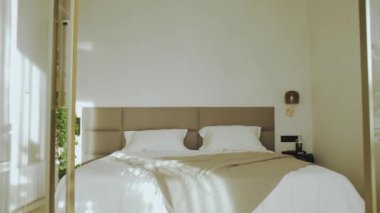 Yatak odası girişinin ön görüntüsü, açık cam kapılar, bej kapaklı yatağa güneş ışınları, yavaş çekim. Yüksek kalite 4k görüntü