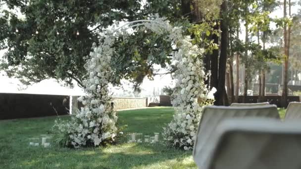 Gros plan belle arche de mariage décorée de fleurs blanches et de verdure dans le parc, pins en arrière-plan, chaises blanches, mariage classique, ralenti. Images 4k de haute qualité