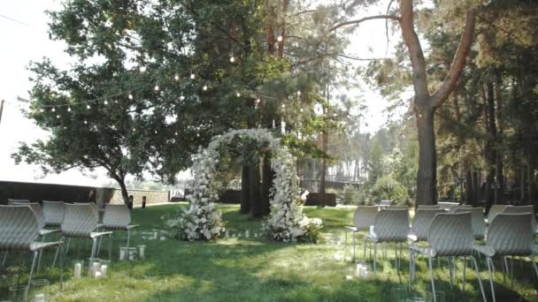 公園の白い花と緑で装飾された美しい結婚式のアーチ 背景の松の木 白い椅子 古典的な結婚式 ゆっくり動き 高品質の4K映像 — ストック動画