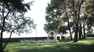 Parkta beyaz çiçekler ve yeşilliklerle süslenmiş güzel bir düğün kemeri, arka planda çam ağaçları, beyaz sandalyeler, çimlerde güneş ışınları, klasik düğün, yavaş çekim. Yüksek kalite 4k görüntü