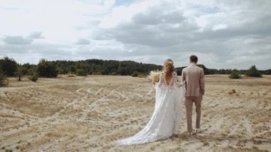 Newlyweds in love walk through the countryside, Bride in boho dress and curly hair and groom in suit goes across the field, slow motion. High quality 4k footage