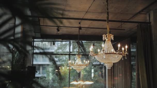 レストランにぶら下がっている美しいシャンデリア 背景に大きな窓 前景にヤシの枝 シャンデリアの多くの透明粒子 スローモーションショット 高品質の4K映像 — ストック動画