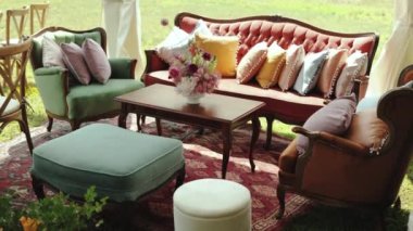 Düğünde misafirlerin dinlenmesi için dekore edilmiş bir yer, ahşap bir masa ve üzerinde güzel bir buket, puf ve üzerinde yastık olan kırmızı antika bir kanepe, yavaş çekim. Yüksek kalite 4k görüntü